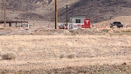 A gated checkpoint featuring a Tesla sign, stop sign, guard booth, floodlight pole, and parked trucks