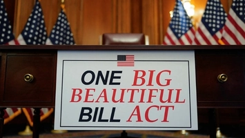 One Big Beautiful Bill Act sign displayed on a wooden desk with American flags arranged in the background