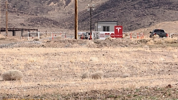 A gated checkpoint featuring a Tesla sign, stop sign, guard booth, floodlight pole, and parked trucks
